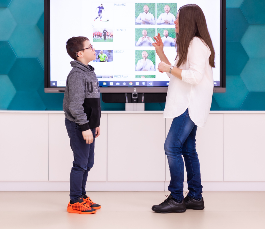 Nauczycielka rozmawiająca z uczniem w języku migowym na tle telewizora z obrazkami dotyczącymi tego języka