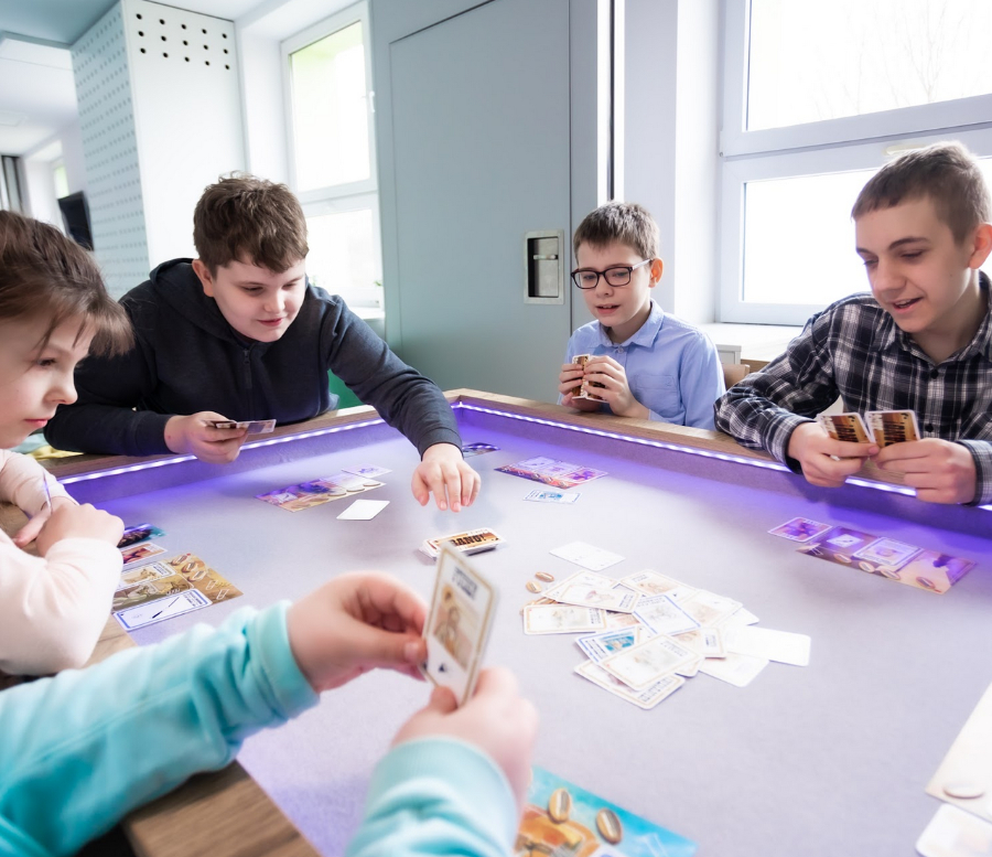 Uczniowie siedzący przy dużym podświetlanym stole, grający w karty